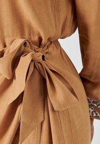 Robe marron en tissu texturé, avec un détail de nœud noué à la taille et un poignet à motif dépassant de la manche.