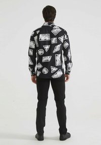 Chemise noire à manches longues avec une variété de motifs géométriques blancs. Associée à un pantalon slim noir et des chaussures noires.
