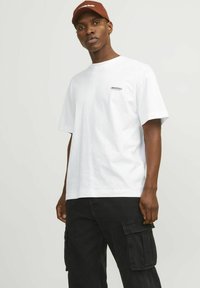 T-shirt bianco in cotone con maniche corte e una stampa sul petto che recita "ORIGINALS". Abbinato a pantaloni cargo neri con tasche laterali.