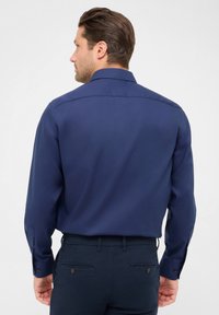 Camicia da uomo blu navy con maniche lunghe, colletto con bottoni, texture liscia e dettaglio sulla spalla. Indossata con pantaloni scuri abbinati.