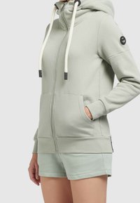 Donna che indossa una felpa con zip verde chiaro, con grandi lacci bianchi e pantaloni corti abbinati, mano nella tasca della felpa, su uno sfondo semplice.