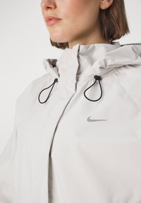 Hellgraue Kapuzenjacke mit hohem Kragen, ausgestattet mit Kordelzügen und einem kleinen grauen Nike-Logo auf der Brust. Glattes, wasserabweisendes Material.