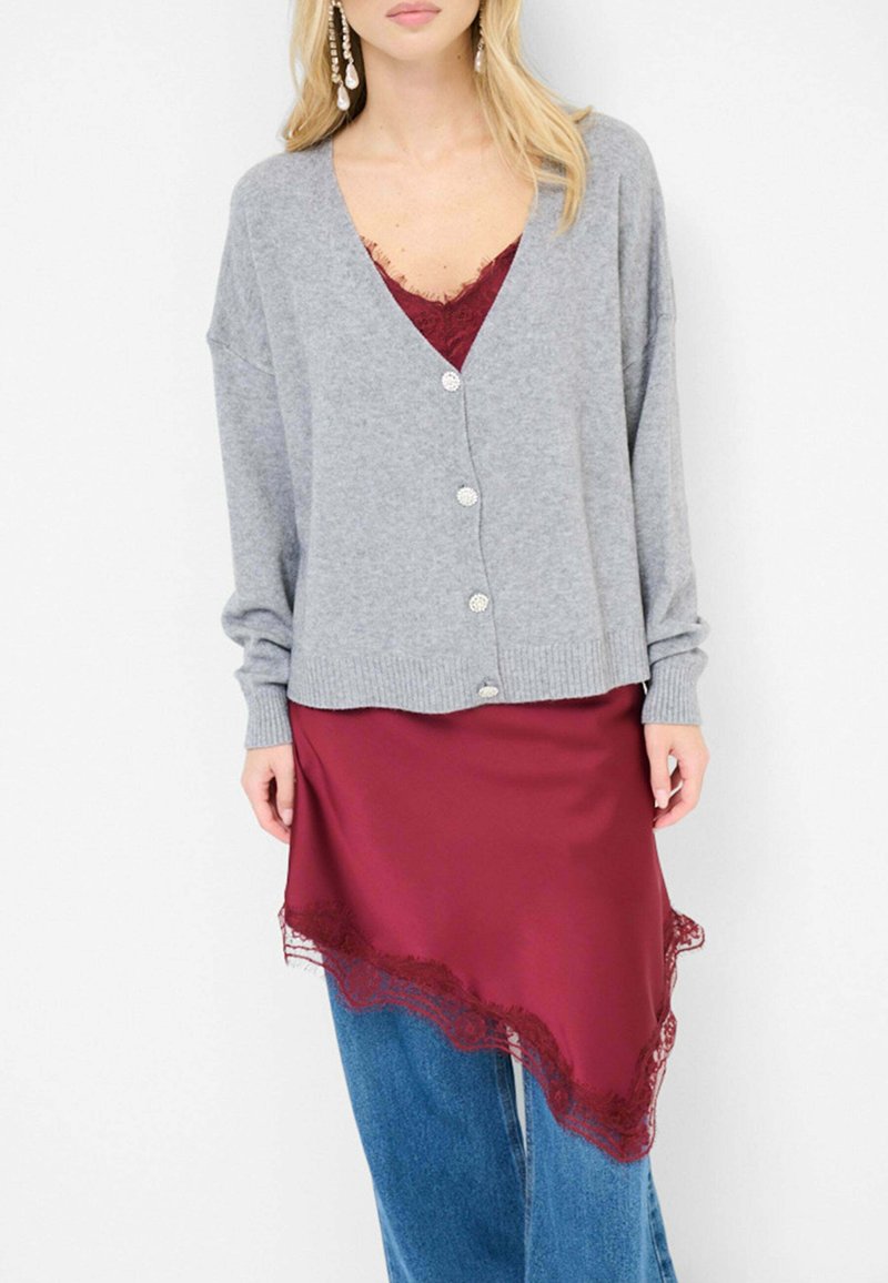Cardigan grigio in maglia con scollatura a V e chiusura con bottoni, indossato su un abito di raso bordeaux con bordi in pizzo, abbinato a jeans larghi blu.