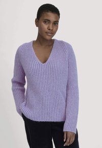 Lilafarbenes Strickpullover mit V-Ausschnitt, geripptem Muster und langen Ärmeln. Weiches, flauschiges Material sorgt für ein gemütliches Aussehen. Kombiniert mit dunklen Hosen.
