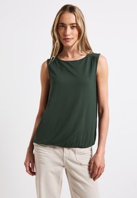 Mörkgrön ärmlös topp med rund halsringning, lätt material, draperade axeldetaljer, matchad med ljusbeiga byxor.