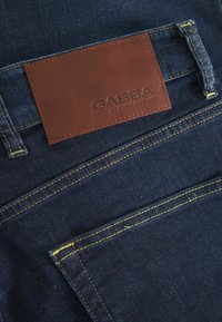 Sötétkék farmernadrág sárga varrással. A hátsó zsebnél található egy barna bőr címke, amelyen a "GABBA" felirat látható.