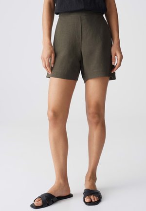 Vrouw draagt olijfgroene shorts met hoge taille, zwart mouwloos topje en zwarte platte sandalen, staand op een effen lichte achtergrond.