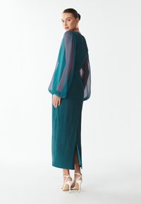 Türkisfarbenes Kleid aus strukturiertem Stoff, mit transparenten langen Ärmeln, seitlichem Schlitz und Rückenschluss. Kombiniert mit weißen High-Heels.