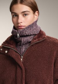 Hnědá fleecová bunda s zapínáním na zip, s vysokým límcem a knoflíkovými detaily, nošená přes fialový pletený rolák.