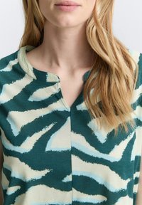 Haut à manches courtes présentant un motif abstrait teal, beige et bleu. Design en V avec une texture douce et des détails de couture subtils.