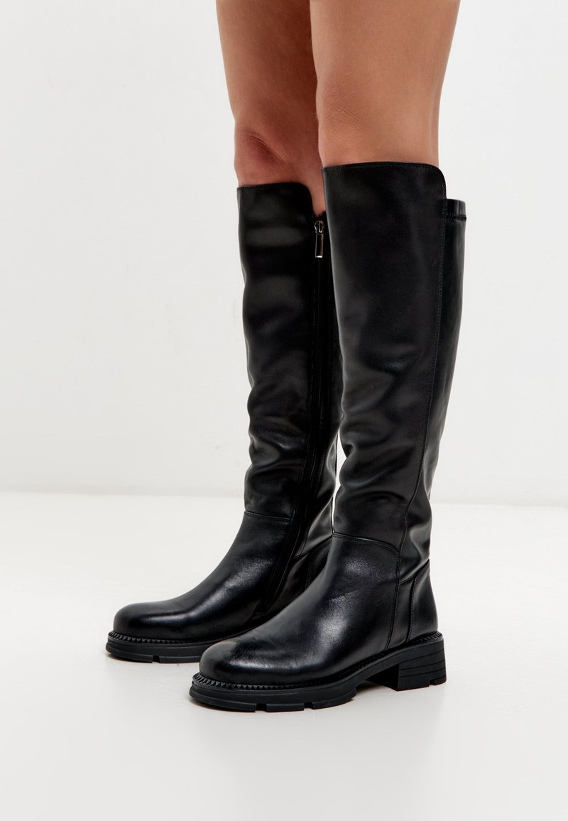 Botas de cuero negro hasta la rodilla con puntera redondeada, cremallera lateral y suela texturizada. Presenta un tacón grueso y una superficie lisa.