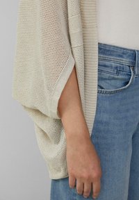 Helle beige Strickjacke mit einem offenen, fließenden Design, das ein strukturiertes Muster und weite Ärmel aufweist. Getragen über einem enganliegenden weißen Oberteil und Jeans.