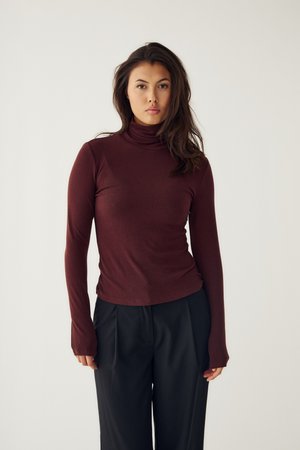 Jeune femme aux longs cheveux foncés portant un col roulé bordeaux et un pantalon noir, debout devant un fond clair uni.