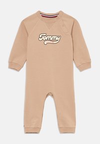 Jumpsuit beige para niños pequeños con mangas largas y puños acanalados, que presenta un prominente logo "Tommy" y cierre a presión en el hombro.