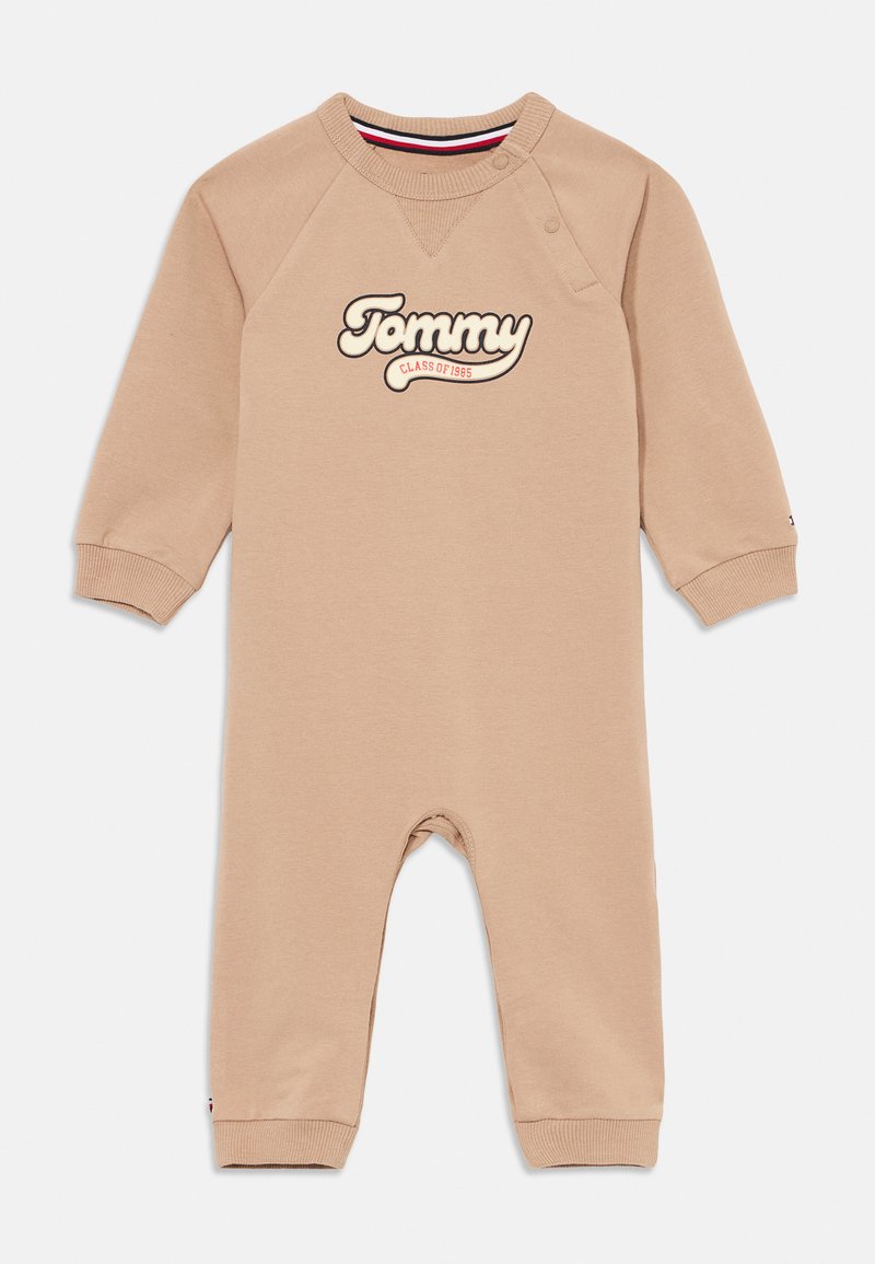 Jumpsuit beige para niños pequeños con mangas largas y puños acanalados, que presenta un prominente logo "Tommy" y cierre a presión en el hombro.