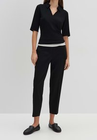 Schwarzes Polohemd mit kurzen Ärmeln, kombiniert mit maßgeschneiderten schwarzen Hosen und flachen schwarzen Schuhen. Glatter Stoff, schlichtes Design, ohne Muster.