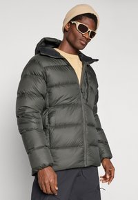 Casaco puffer verde escuro com fecho à frente, capuz e textura acolchoada. Combinado com um gorro de malha bege e óculos de sol.