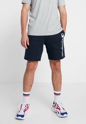 Mørkeblå shorts lavet af blødt stof, med hvidt "Champion"-logo på siden. Kombineret med en lysegrå t-shirt og farverige sneakers.