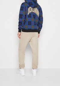 Felpa con cappuccio a quadri blu con accenti verdi e rossi, con la scritta "JORDAN" in beige, abbinata a pantaloni beige e sneakers bianche.
