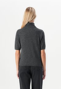 En blond kvinne som vender bort, iført en mørk grå kortermet høyhalset genser og svarte høyreiste jeans mot en hvit bakgrunn.