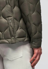 Giacca trapuntata verde scuro con bottoni a pressione, indossata su pantaloni grigi chiari, che mostra dettagli di cucitura e una mano appoggiata vicino alla tasca.