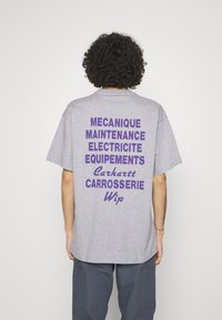 Person med krøllet hår iført en grå T-shirt med lilla franske ord om mekanik og vedligeholdelse på ryggen, kombineret med mørke bukser.