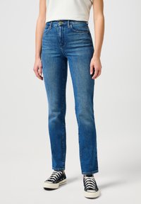 Jean en denim bleu taille haute avec une coupe droite, un léger délavage et un design classique à cinq poches. Portés avec des baskets noires.