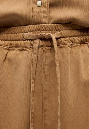 Primer plano de pantalones de tela marrón con cintura elástica y cordón atado en la parte central delantera.