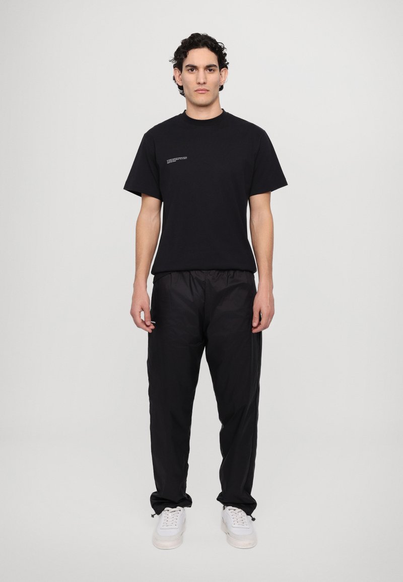 Tricou negru din bumbac cu guler rotund și text mic alb pe piept, asortat cu pantaloni negri ușori și adidași albi.