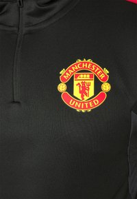 Gros plan sur une veste de sport noire arborant le blason rouge et jaune du club de football Manchester United sur la poitrine.