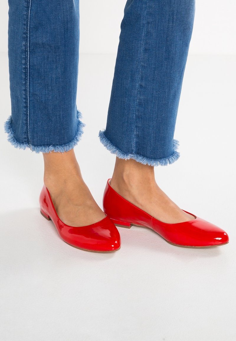Czerwone baleriny z lakierowanej skóry ze spiczastym noskiem, niskim obcasem i gładką powierzchnią, zestawione z niebieskimi dżinsami z postrzępionymi mankietami.