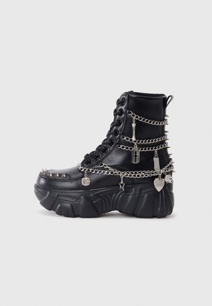 Černé kotníkové boty vyrobené z umělého materiálu, s masivní podrážkou, ozdobené stříbrnými řetězy, hroty a kovovými přívěsky.