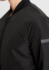 Veste bomber noire en tissu léger et brillant. Dotée d'un col côtelé, d'une fermeture éclair et d'une petite étiquette avec le logo de la marque sur la manche.