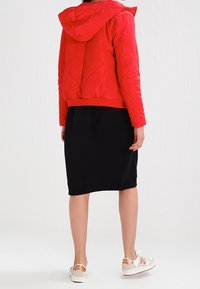 Rode gewatteerde pufferjas met capuchon, elastische manchetten en tailleband, gedragen over een zwarte knielange rok en witte sneakers.