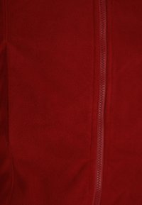 Raudonas fliso švarkas su viso ilgio užtrauktuku ir minkštu, tekstūruotu paviršiumi. Nėra matomų kišenių ar papildomų puošmenų.