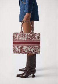 Canvas indkøbstaske med blomsterudsmykning i rødt på en neutral baggrund. Har to håndtag og en kontrasterende vandret stribe.