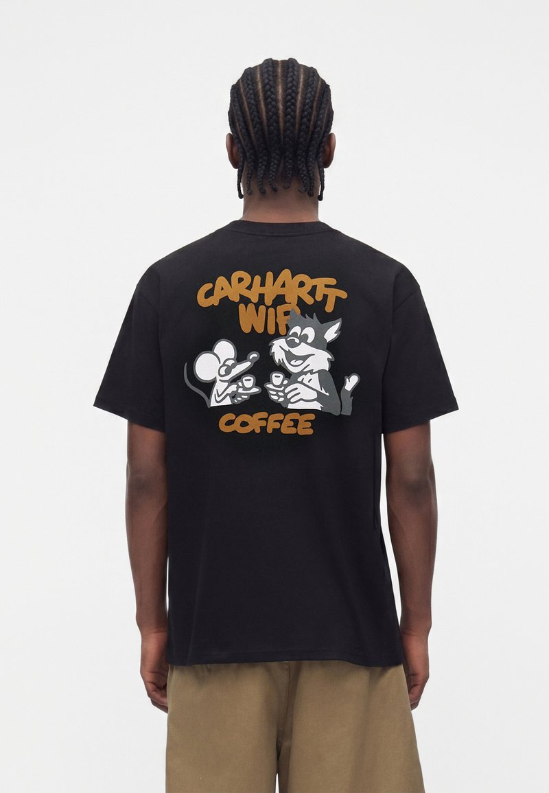 Person mit geflochtenem Haar, die ein schwarzes T-Shirt trägt, auf dem eine Cartoon-Maus und ein Wolf zusammen Kaffee trinken, mit dem Schriftzug "Carhartt WIP Coffee" auf dem Rücken.