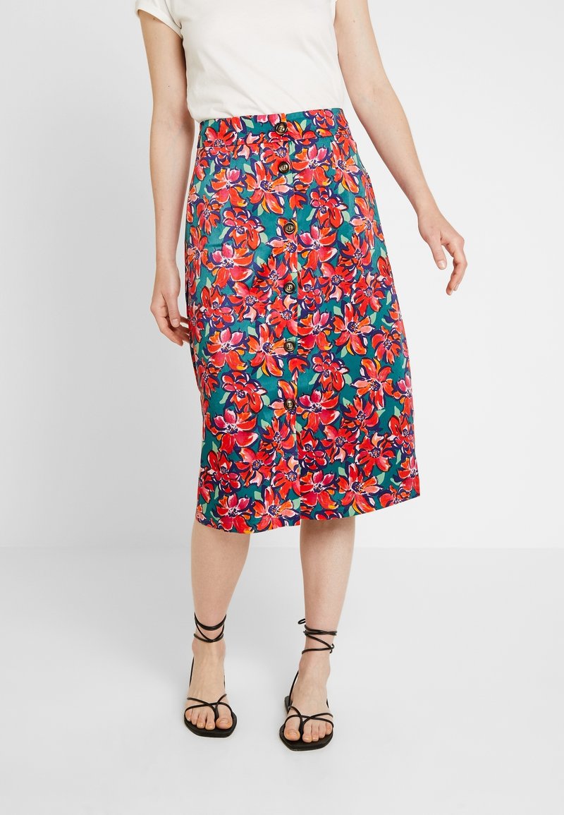 Femme portant une jupe longueur genou à boutons sur le devant, imprimé floral rouge vif et rose sur fond turquoise, et sandales noires à brides.