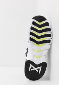 Sort og hvid sneakersål med gule accenter og teksturerede grip-mønstre, placeret lodret mod en hvid hjørnevæg.