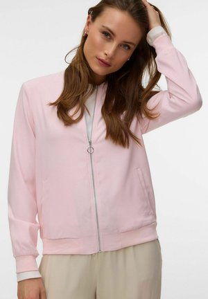 Jonge vrouw met lang bruin haar die een lichtroze jas met rits en beige broek draagt, poseert met één hand op haar hoofd.