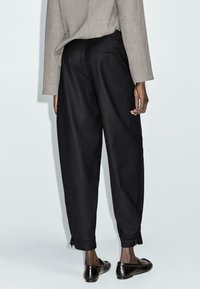 Black tapered trousers made of textured fabric, featuring zip accents at the cuffs and tailored back pockets. Worn with a neutral top.