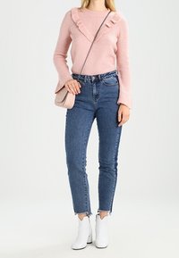 Femme portant un pull à volants rose, un jean taille haute bleu, des bottines blanches et tenant un sac bandoulière rose pâle.
