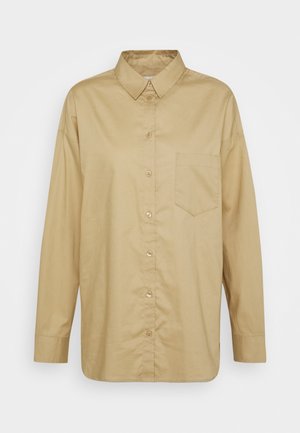 Chemise beige à manches longues avec boutons, col et une poche poitrine unique, présentée sur un fond clair uni.