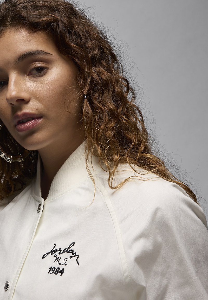 Veste bomber blanche avec logo "Jordan" brodé et "1984". Le tissu semble lisse avec un léger éclat, et une fermeture à boutons-pression.