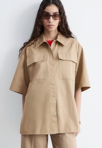 Femme aux longs cheveux bouclés portant une chemise marron oversize à manches courtes et un pantalon assorti, des lunettes de soleil teintées marron et des boucles d'oreilles créoles.
