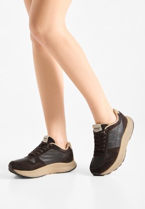 Piernas con zapatillas negras y beige con lados texturizados, de pie sobre un suelo blanco frente a un fondo liso.