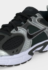 Μαύρο και σκουρόγρινο παπούτσι Nike για τρέξιμο με ασημί επιστρώσεις, μαύρα κορδόνια, αναπνεύσιμα πλέγματα και λευκή σόλα με μαύρες λεπτομέρειες στο πέλμα.