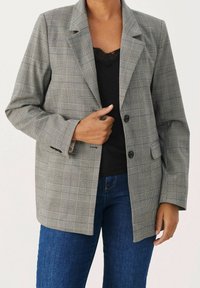 Blazer à carreaux gris avec revers, deux boutons noirs et poches frontales. Porté sur un haut noir et assorti à un jean bleu.
