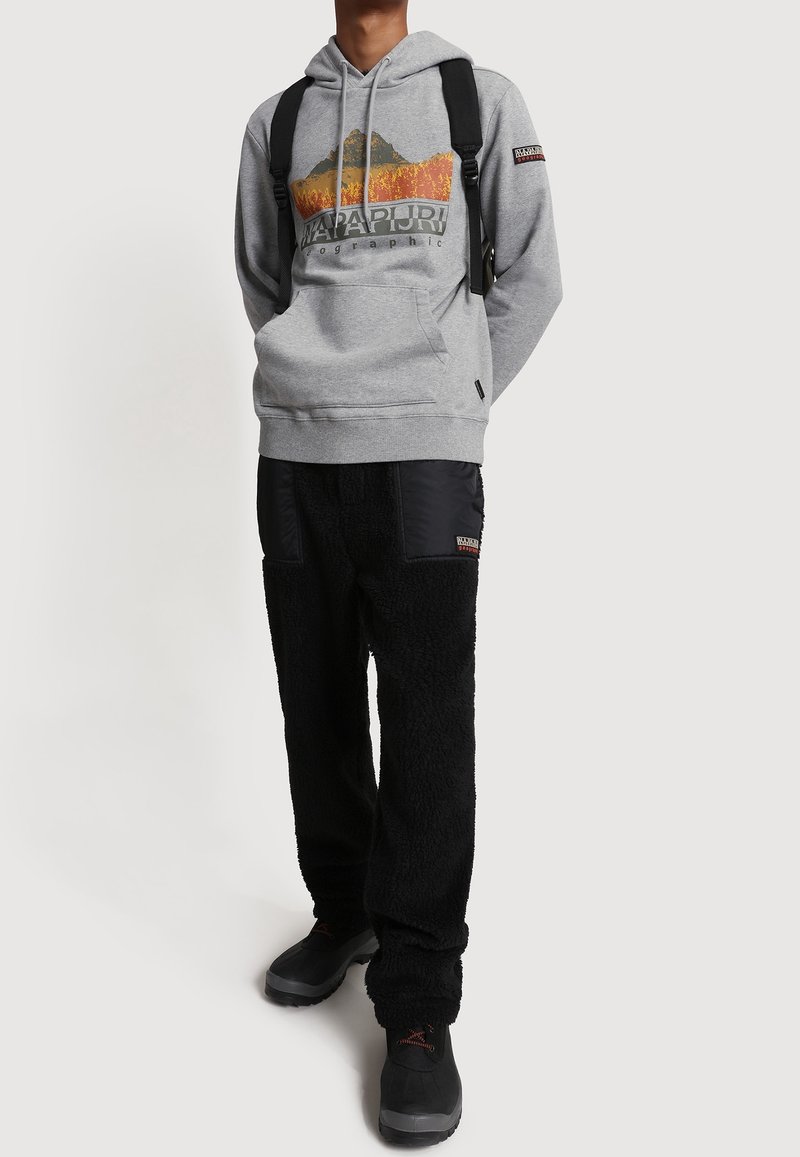Personne portant un sweat à capuche gris avec un motif de montagne, un pantalon noir texturé avec des patchs, des bottes noires et un sac à dos noir.