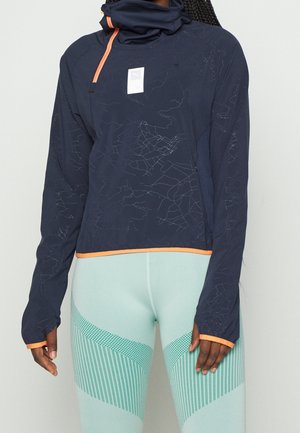 Kvinde iført navyblå atletisk langærmet top med orange kant og lyseblå mønstrede leggings, stående mod en lys baggrund.